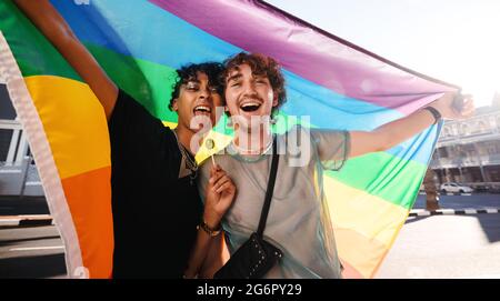 Non-conforming young men celebrating gay pride. Two young gay men smiling cheerfully while raising a rainbow pride flag. Two members of the LGBTQ+ com Stock Photo