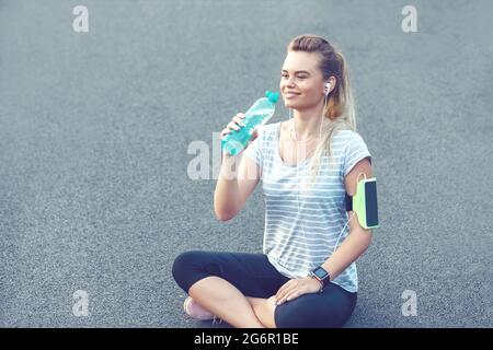 Happy young sporty woman drinking water from bottle while taking a break after running workout Stock Photo