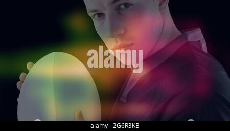 Composition of glowing yellow, blue and red blur over male rugby player holding rugby ball, on black Stock Photo