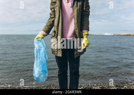 A female volunteer holds a bag of garbage in her hands, collecting garbage on a dirty beach. The problem of environmental pollution Stock Photo