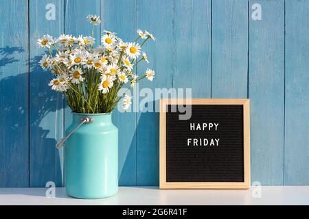 Happy Friday words on black letter board and bouquet of chamomile flowers on table against blue wooden wall. Concept Hello Friday. Stock Photo