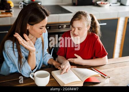 White woman helping her daughter with down syndrome reading book at home Stock Photo