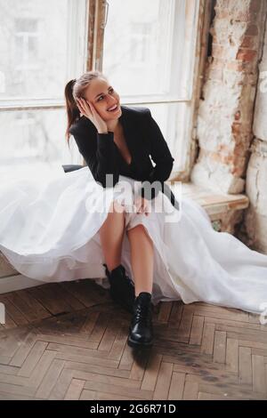 Full body cheerful female in trendy dress and jacket smiling and looking away while sitting near window in loft room Stock Photo