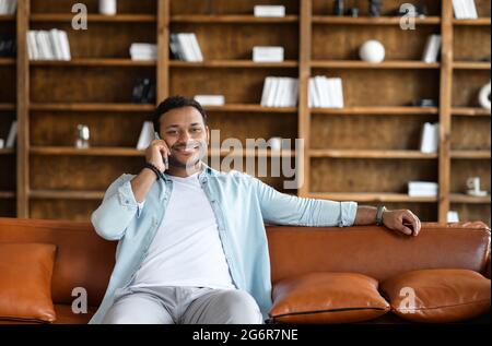 Cheerful carefree young indian hipster guy is chilling at home, sitting on the comfy sofa in the modern living room, holding a mobile phone, talking with the friend, colleague, family Stock Photo