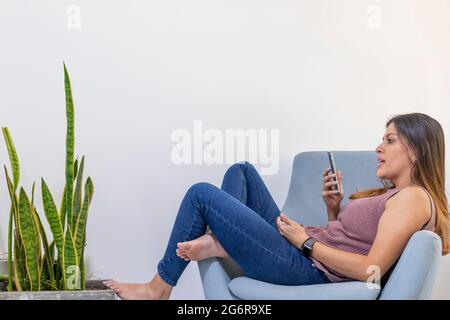 young latina woman in a chair recording a voice message in smart phone. Stock Photo