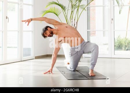 Sporty athletic man doing yoga mobility exercises with a forward lunge side stretch to strengthen and tone his muscles in a high key gym with copyspac Stock Photo