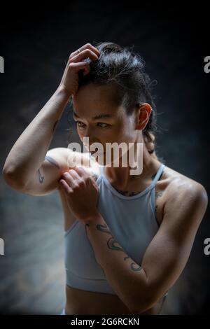 A young woman with tattoos in gymwear, she is holding one arm on top of her head and the other is raised. She is taking a break from exercising. Stock Photo