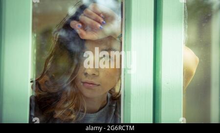 A young woman, photographed through the window. She is resting on her raised arm against the window. Stock Photo