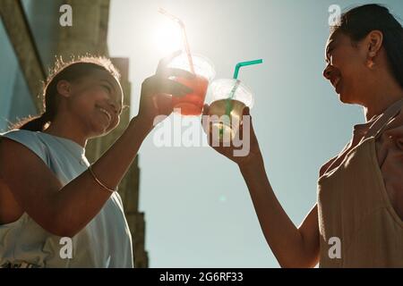 Low angle portrait of two young women clinking cocktail glasses in Summer against sunlit blue sky Stock Photo