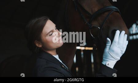 Horsewoman touching the face of her seal brown horse. Girl wearing gloves and dressed in a black coat. Looking at her horse with love. Horse stable. Love for horses. Stock Photo