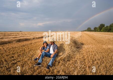 Father and son are resting after successful harvest. Rainbow in the sky behind them. Stock Photo