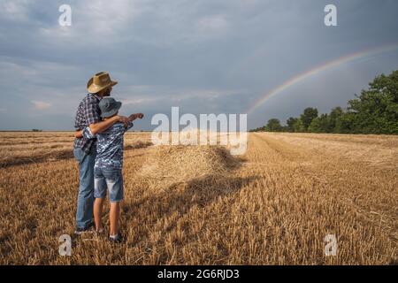 Father and son are standing in their wheat field after successful harvest. They are looking at rainbow in the sky. Stock Photo