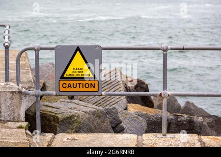 A sign in front of rocks at the coast reading caution please do not climb on rocks Stock Photo