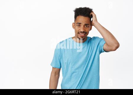 Awkward worried african american man scratch head guilty, furrow eyebrows and looking confused, puzzled to answer question, standing against white Stock Photo