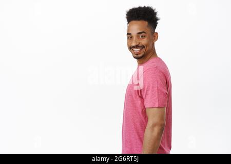 Portrait of happy and confident african american guy, standing in profile, turn head at camera with big cheerful smile onhis face, standing in pink t Stock Photo