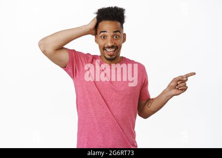 Surprised handsome african american man, pointing finger right at sale banner, touching head awkward or shy, recommend something, showing Stock Photo