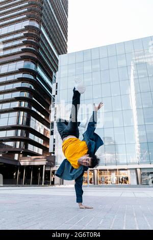 Talented young man with casual clothes doing break moves in the street against big modern buildings in the city. Stock Photo