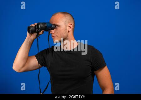Young man over isolated blue wall and looking in the distance with binoculars. Exploration concept Stock Photo