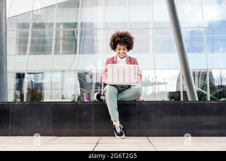 Business woman with afro hair wearing formal clothes sitting outdoors in business area and using her computer. Stock Photo