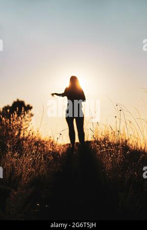 Silhouette of young girl walking at mountain top against sunset. Woman outdoors in nature walking, strolling, wandering at the countryside, forest or Stock Photo