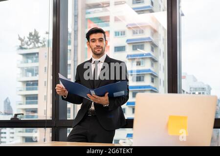Caucasian smart executive businessman standing holding document folder looking through laptop in the office Stock Photo