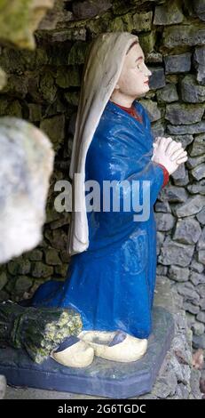 Statue of a peasant woman in a blue dress kneeling before Mother Mary. She looks like a nun. Next to her are a pair of wooden clogs Stock Photo