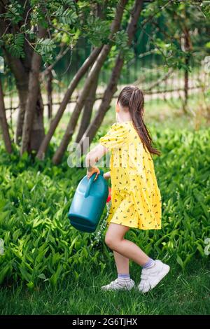 a full-length portrait of a girl in a yellow dress watering lilies of the valley from a blue watering can and helping a family in the backyard on a Stock Photo