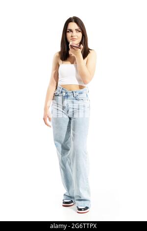 Displeased skeptical young gen Z stylish casual woman talking on cell phone speaker. Full body portrait isolated on white background Stock Photo