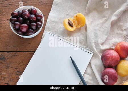 Red and yellow peaches are lying on a linen tablecloth. On a red wooden table, red cherries are lying in a white bowl. Notebook with a pen is lying on Stock Photo