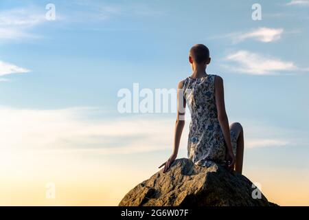 slender girl with a short haircut in a summer dress sits on a rock with her back to the viewer against the sky Stock Photo