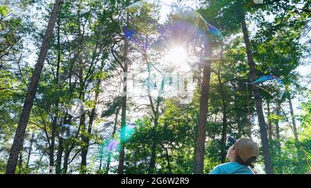 The boy looks at the huge rainbow soap bubbles blown out by the animator in the amusement park. Beautiful soap bubbles on the background of trees and Stock Photo