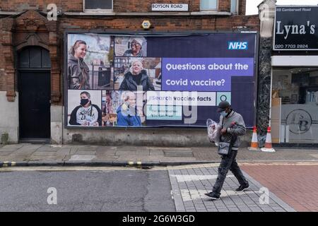 A south Londoner wearing a face covering walks past a governement NHS (national Health Service) billboard giving a link for official information relating to Covid vaccine facts, an attempt to counter misinformation and conspiracy theories to the public in Lambeth - a borough that has seen high infection rates of Covid and Delta variants, on 6th July 2021, in Camberwell, London, England. Stock Photo