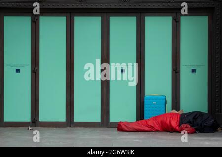 An anonymous homeless person lies on the ground next to a suitcase in a red sleeping bag, in the doorway of the former high street retailer, Top Shop at Oxford Circus in the West End, on 8th July 2021, in London, England. Stock Photo