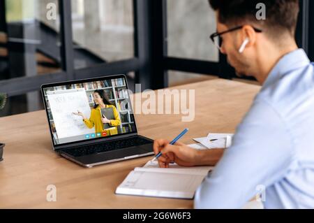 E-learning concept. Clever caucasian male student gains knowledge watching lesson online and studying distantly. Guy takes notes during webinar, listening lecture from female teacher on video call Stock Photo
