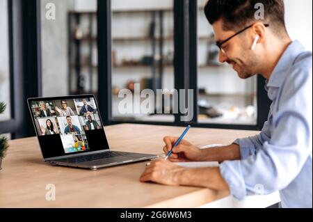 Video conference with teacher and students. Clever caucasian guy watching lesson online and studying distantly. Young man takes notes, listening lecture from female teacher on video call Stock Photo