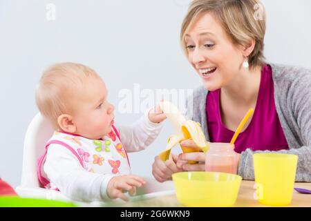 Portrait of a happy mother giving a fresh and nutritious banana to her cute 6 month old baby girl, while starting to eat solid food at home Stock Photo