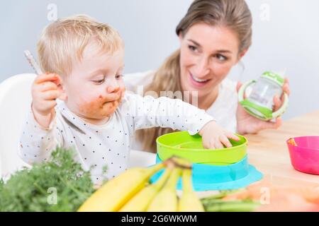 Portrait of a cute and funny baby girl sitting on high chair, while eating with her hands solid food from a plastic bowl next to her dedicated mother Stock Photo