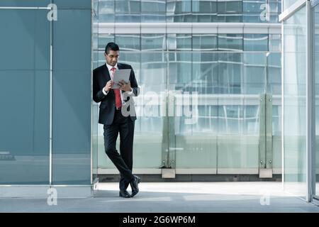 Indian businessman using a tablet PC while leaning on the wall of a modern office building indoors Stock Photo