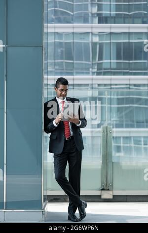 Indian businessman using a tablet PC while leaning on the wall of a modern office building indoors Stock Photo