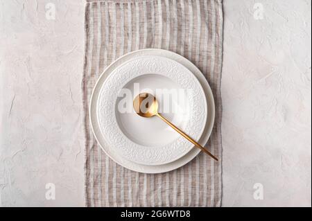 Empty white plate and golden spoon on napkin. Kitchen utensils set on light gray table. Top view flat lay with copy space Stock Photo