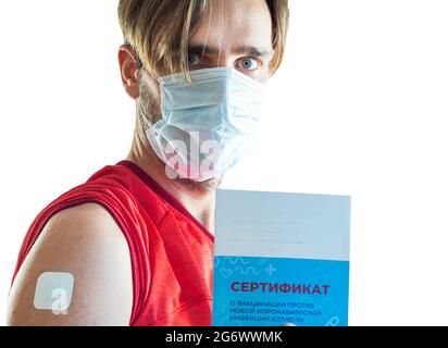 A man in a medical mask holds a vaccination certificate in his hand. The injection site on the shoulder is sealed with a plaster. Translation: certifi Stock Photo