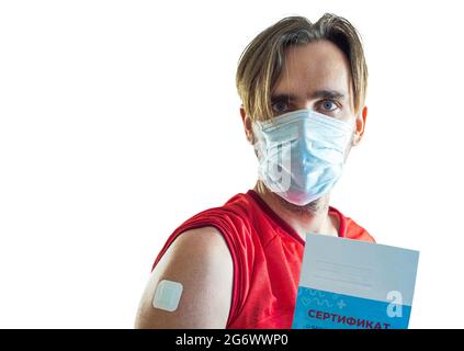 A man with a plaster pasted on his shoulder and a medical mask holds a vaccination certificate in his hand. Translation: certificate of vaccination ag Stock Photo