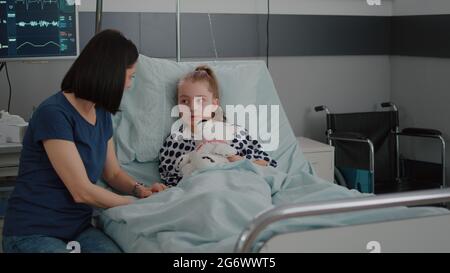 Mother explaining medication treatment to sick daughter during recovery examination in hospital ward. Hospitalized girl child resting in bed wearing oxygen nasal tube suffering disease infection Stock Photo