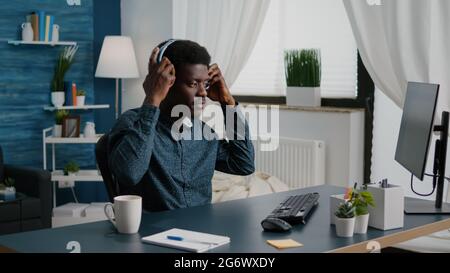 Black freelancer of african ethnicity putting headphones on while working from home, remote worker computer user in home office typing on PC listening music. Concentrated guy enjoying music Stock Photo