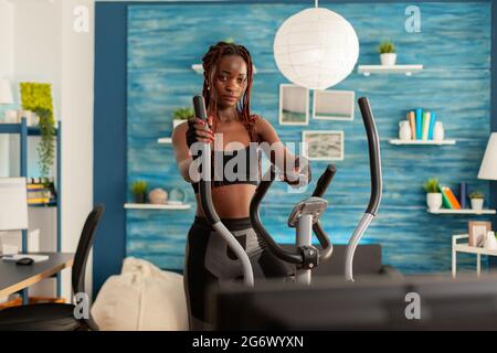 Strong athletic fit black woman doing cross training exercising in home living room using modern eliptic cardio equipment. exercising and changing tv channels. Stock Photo