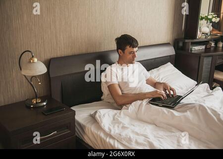 A middle-aged man is sitting in bed in his bedroom and working on a laptop. The concept of remote work Stock Photo