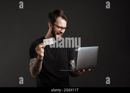 Bearded happy man laughing while showing credit card and using laptop isolated over black background Stock Photo
