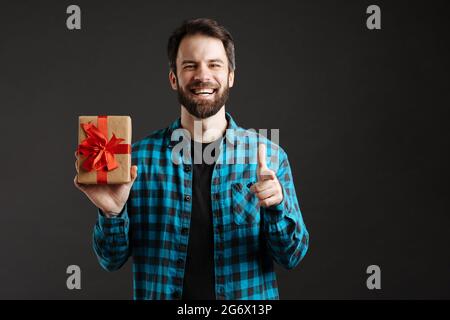 Bearded happy man pointing finger at camera while showing gift box isolated over black background Stock Photo