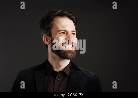 Bearded happy man in black jacket smiling and looking aside isolated over black background Stock Photo