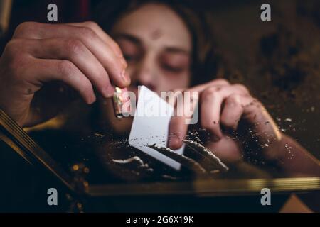 Close-up low-angle view of the hands of an addicted young man snorting powdered cocaine with a rolled banknote at home Stock Photo